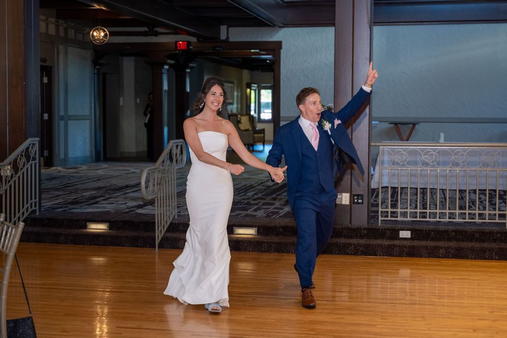 Bride in white gown and groom in navy suit make grand entrance holding hands on dance floor at wedding reception.