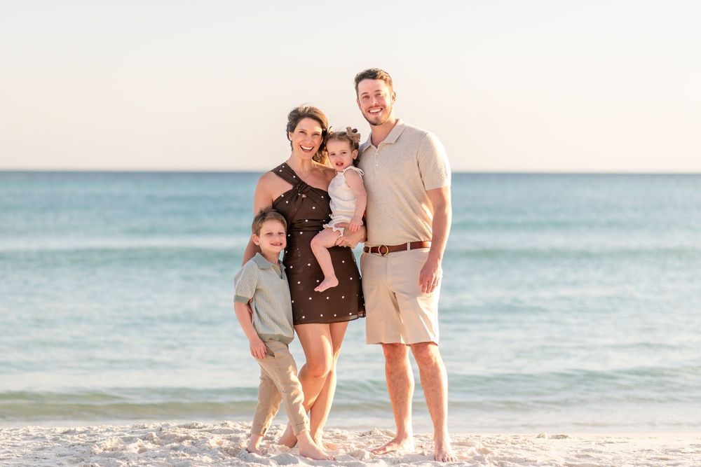Family of four smiling together on a sunny beach with calm turquoise water in the background.