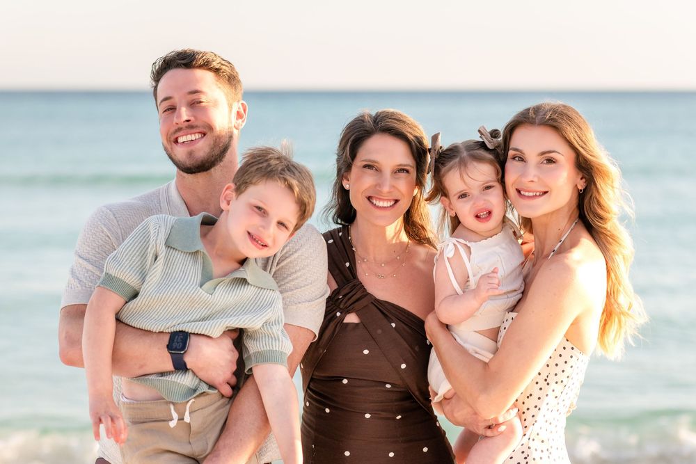 Happy family of five posing together on a sunny beach, with two young children held by their parents and grandparent.
