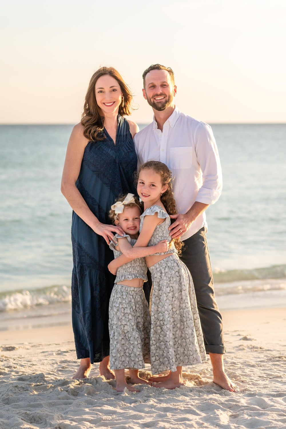 Happy family of four posing on a sunny beach, parents and two daughters in matching dresses smiling by the ocean.