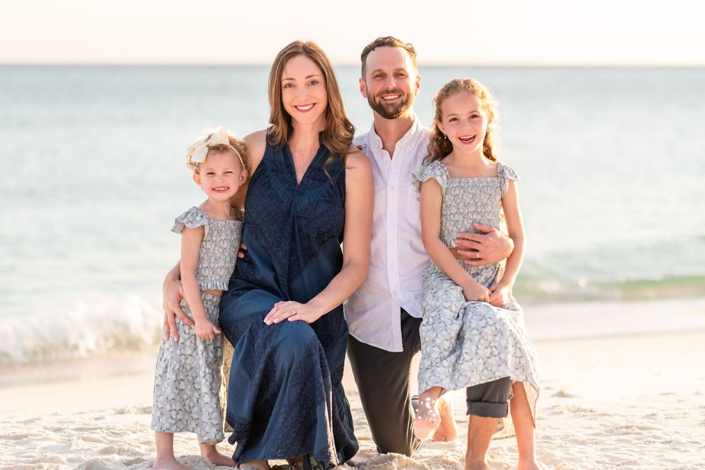 Family of four smiling together on a sunny beach, parents with two young daughters in matching floral dresses.