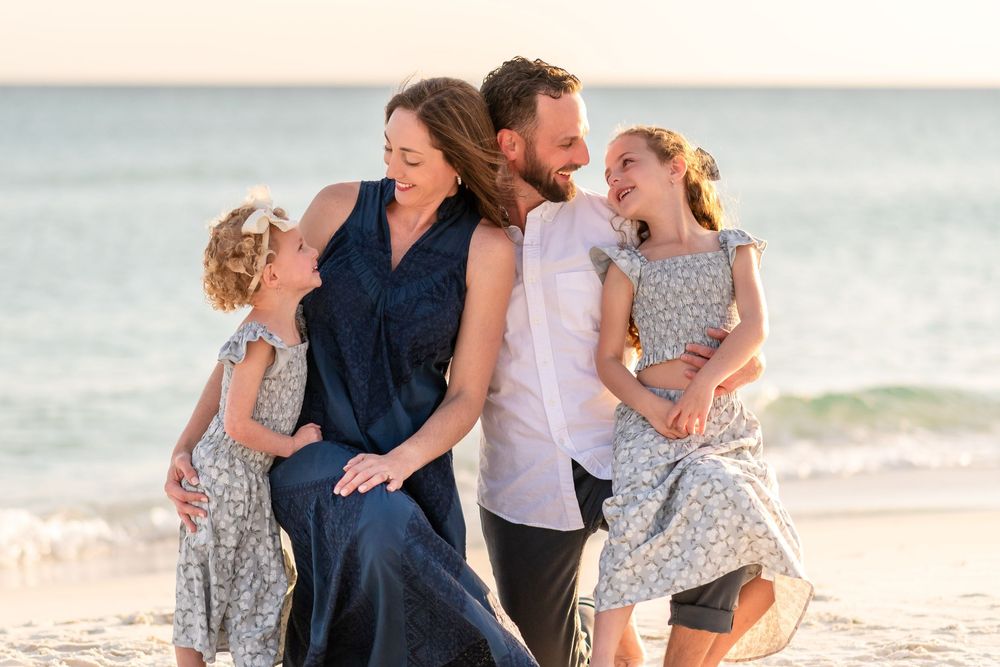 Happy family of four laughing together on a sandy beach at sunset, parents holding two young daughters.