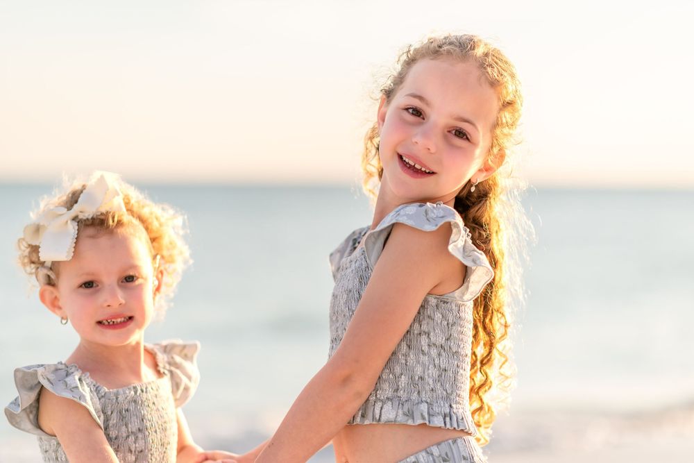 Two young sisters in matching silver dresses smile together on a sunny beach, the older girl holding her little sister's hands.