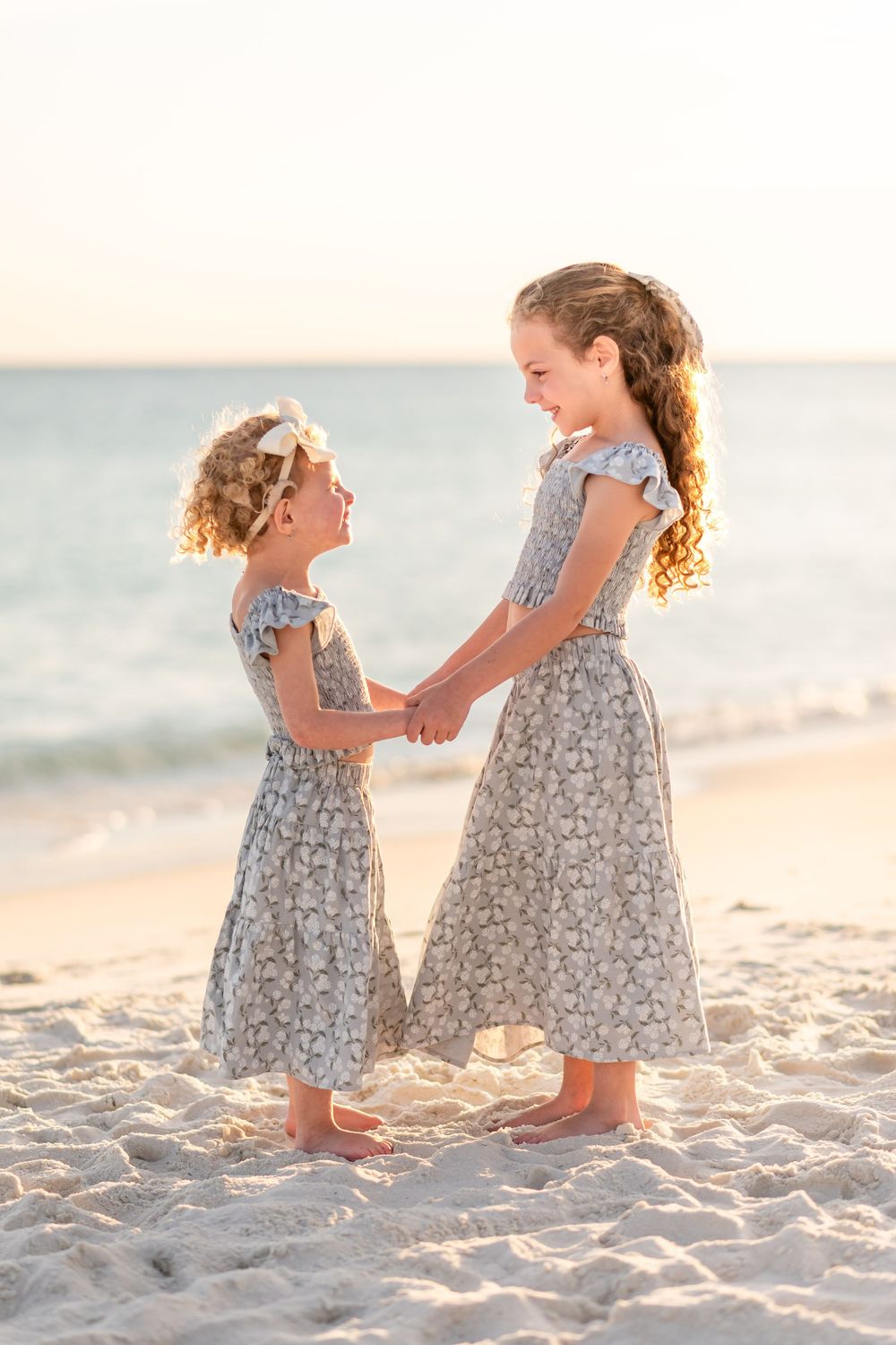 Two young sisters in matching floral dresses hold hands smiling on a sandy beach at sunset.