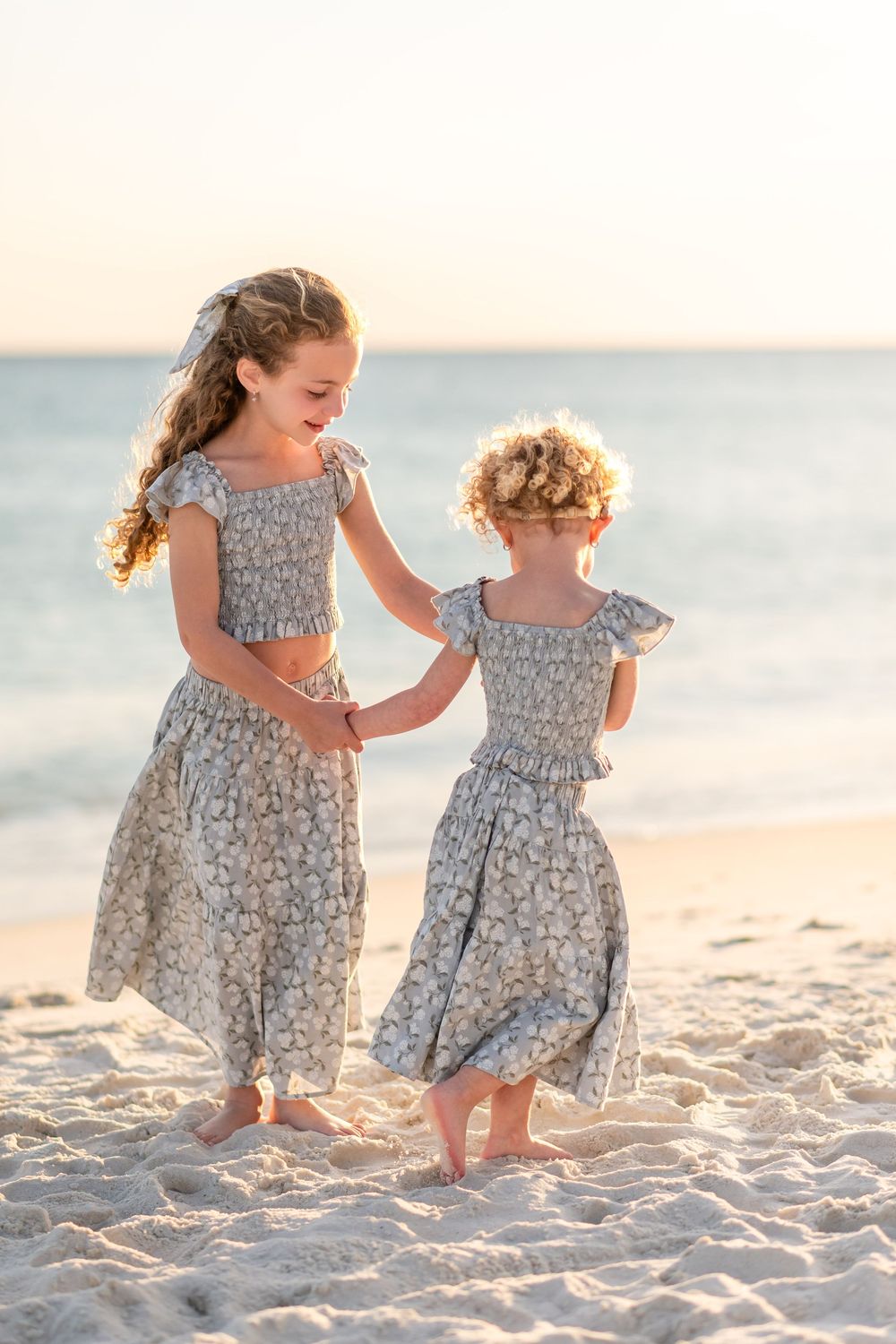 Two young sisters in matching floral crop top and skirt sets holding hands and dancing on a sunny beach.
