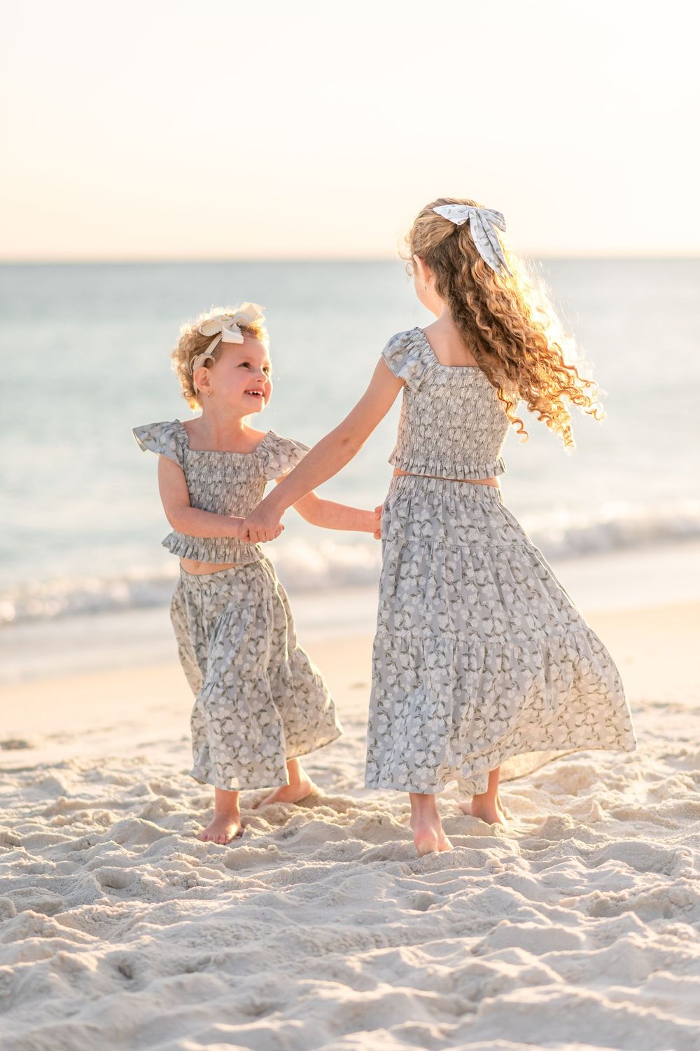 Two young girls in matching floral dresses hold hands and dance on a sandy beach at sunset.