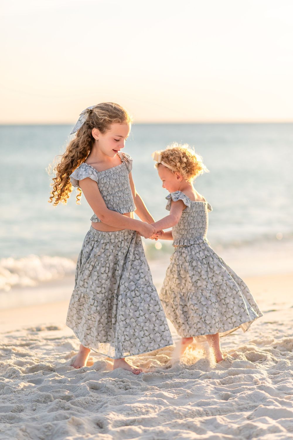 Two young sisters in matching floral dresses hold hands and dance on a sandy beach at sunset.