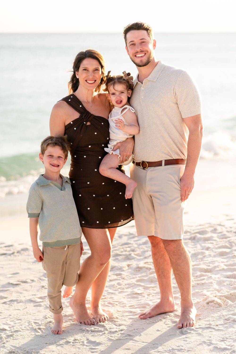 Family of four smiling together on a sandy beach at sunset, parents with two young children in coordinated outfits.