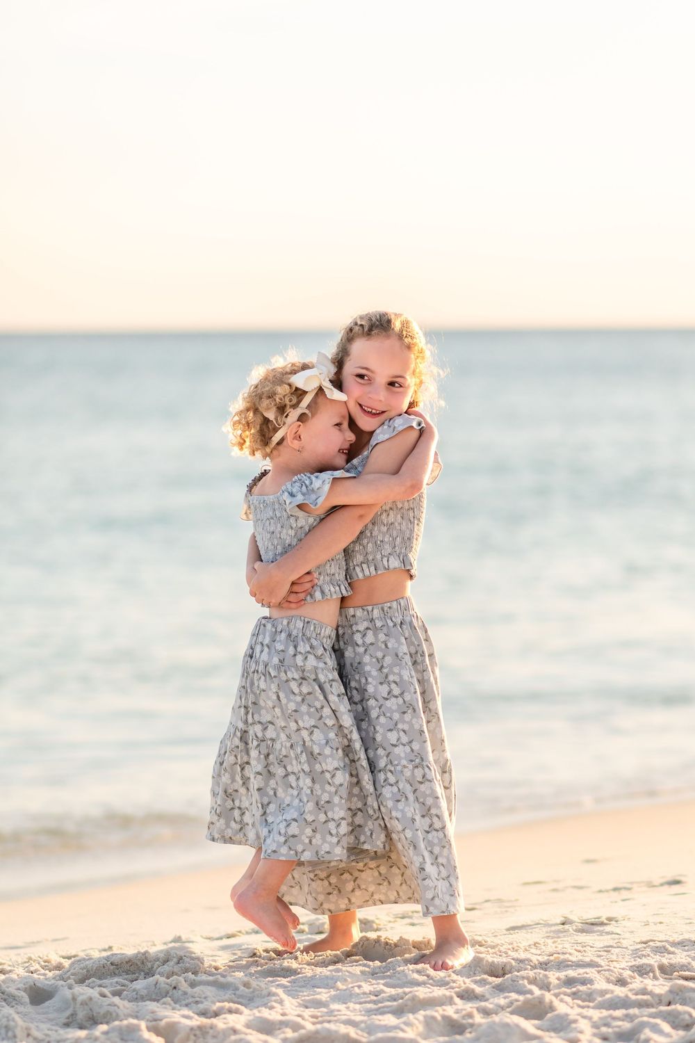 Two girls in matching floral dresses hugging on a sandy beach at sunset.