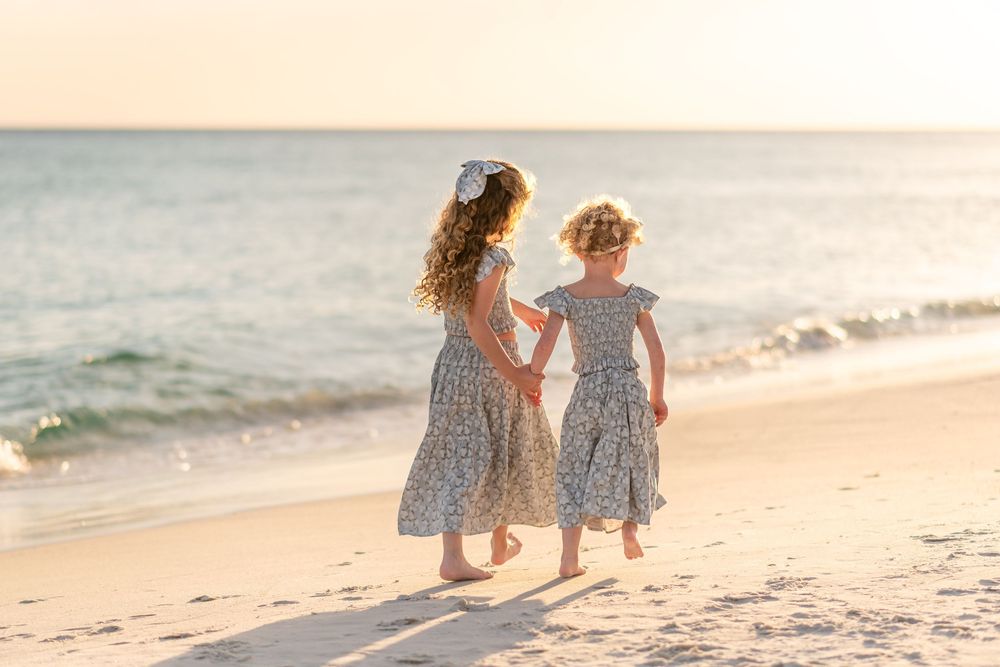 Two young girls in matching floral dresses hold hands walking barefoot on a sandy beach at golden sunset.