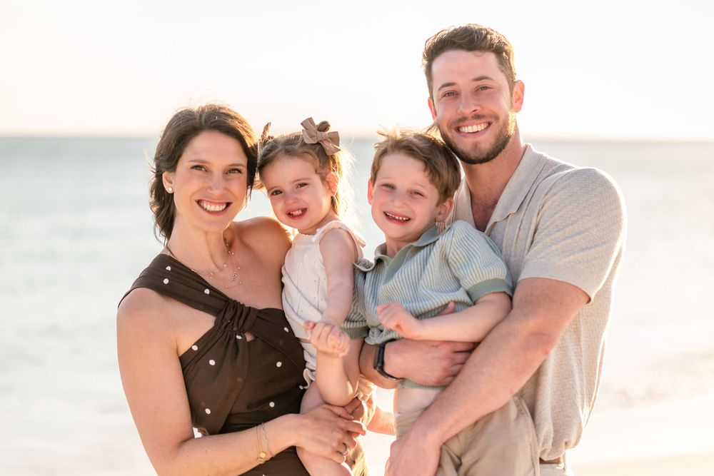 Happy family of four smiling together on a beach at sunset, parents holding two young children.