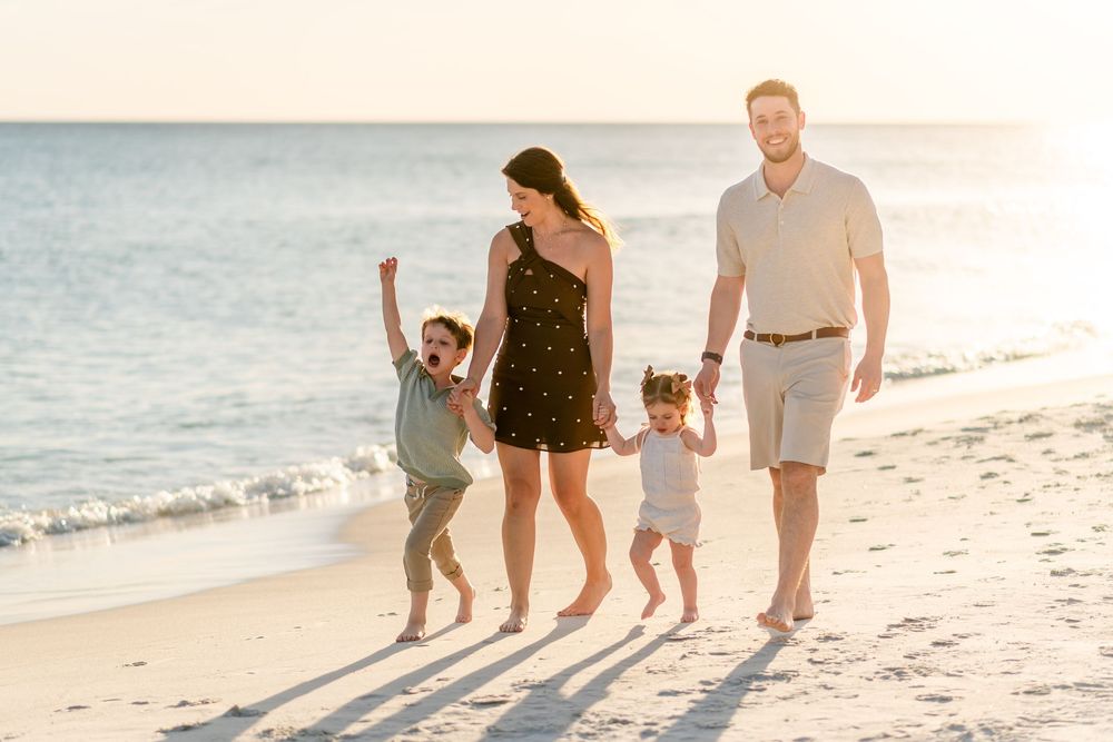 Happy family of four walking on a sunny beach, parents swinging children by their hands near ocean waves.