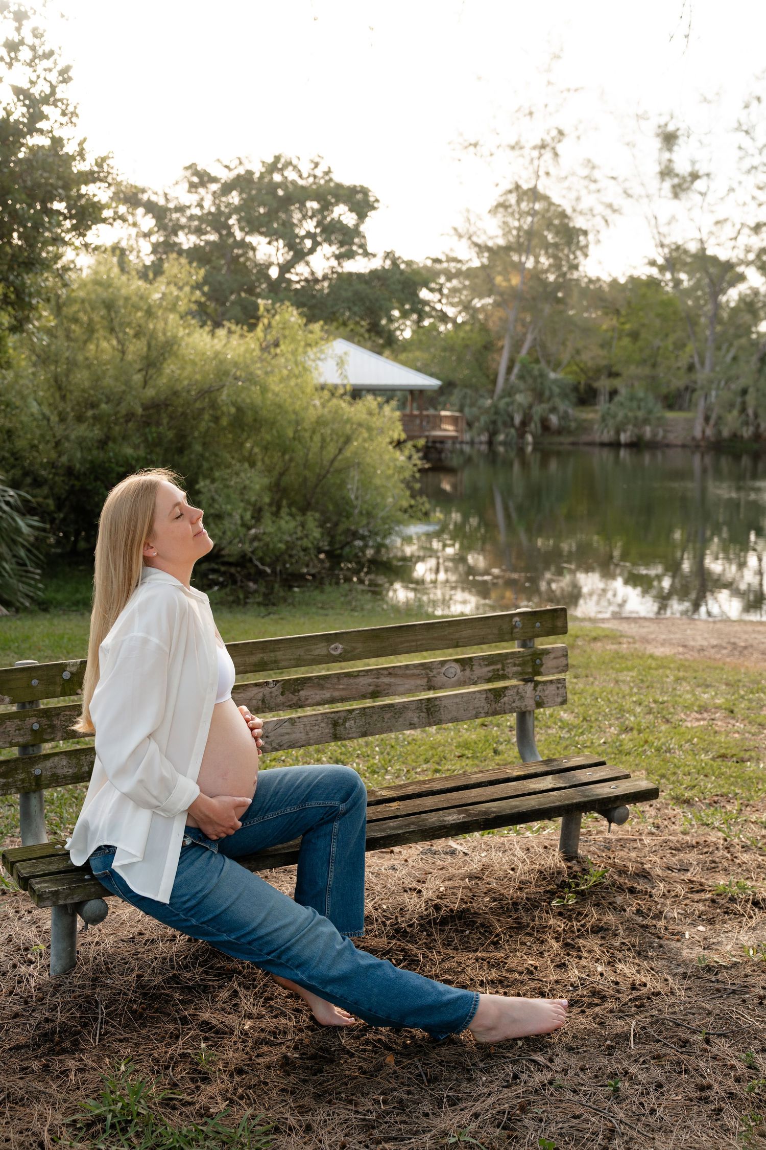 Pregnant woman sitting on a park bench by a pond, wearing white shirt and jeans, enjoying the outdoors.