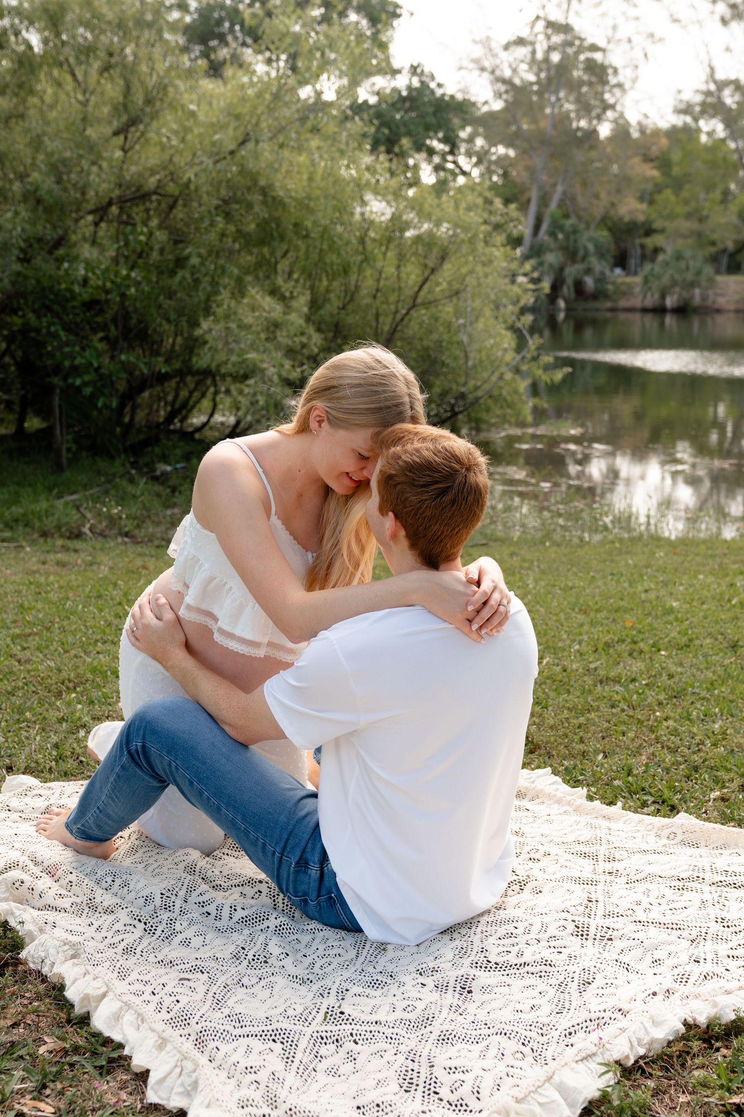 Couple sitting on lace blanket by pond in park, woman in white top and jeans embracing man in white shirt.