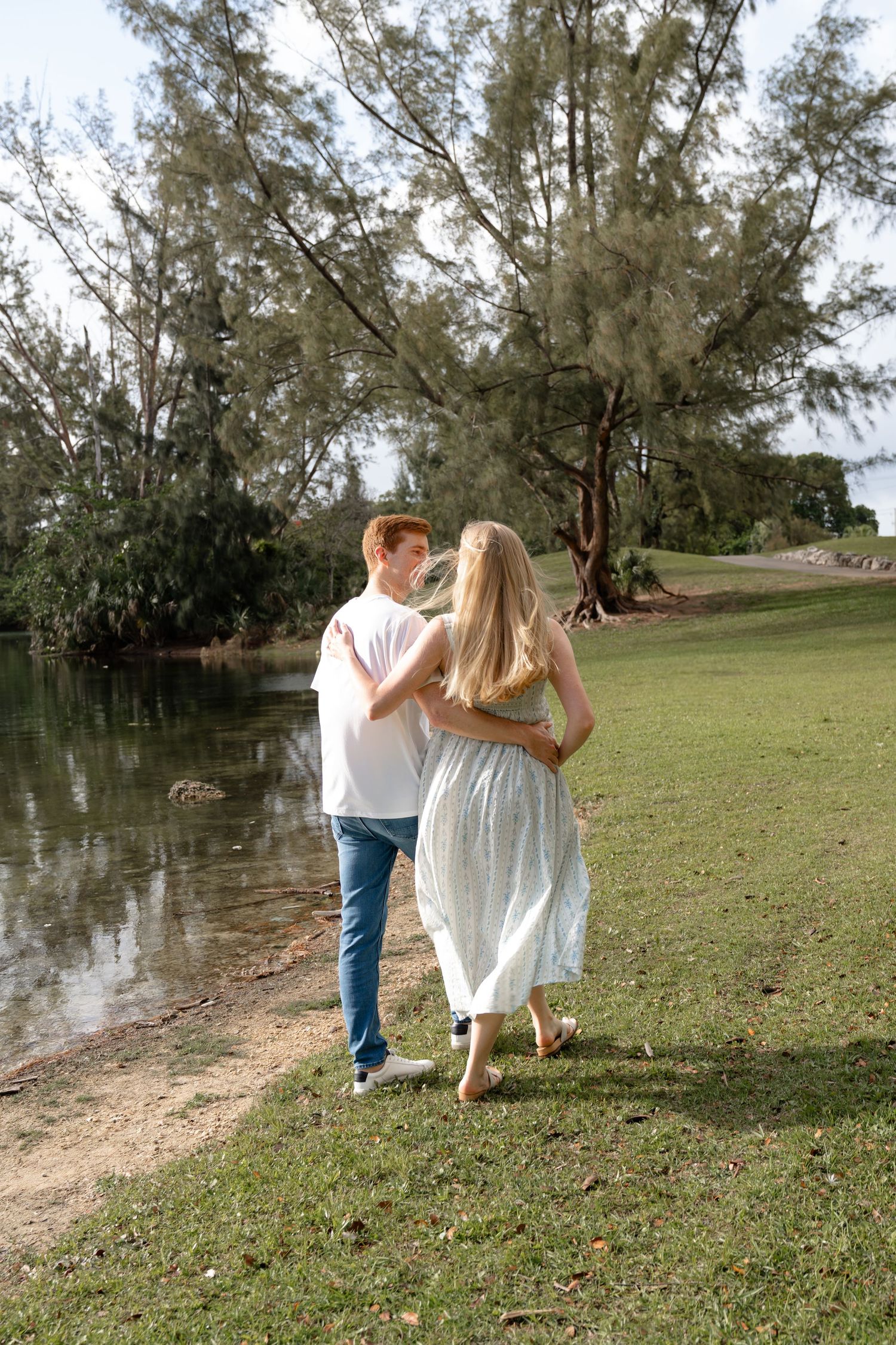 A couple walks hand-in-hand along a serene lakeside path surrounded by lush green trees on a sunny day.