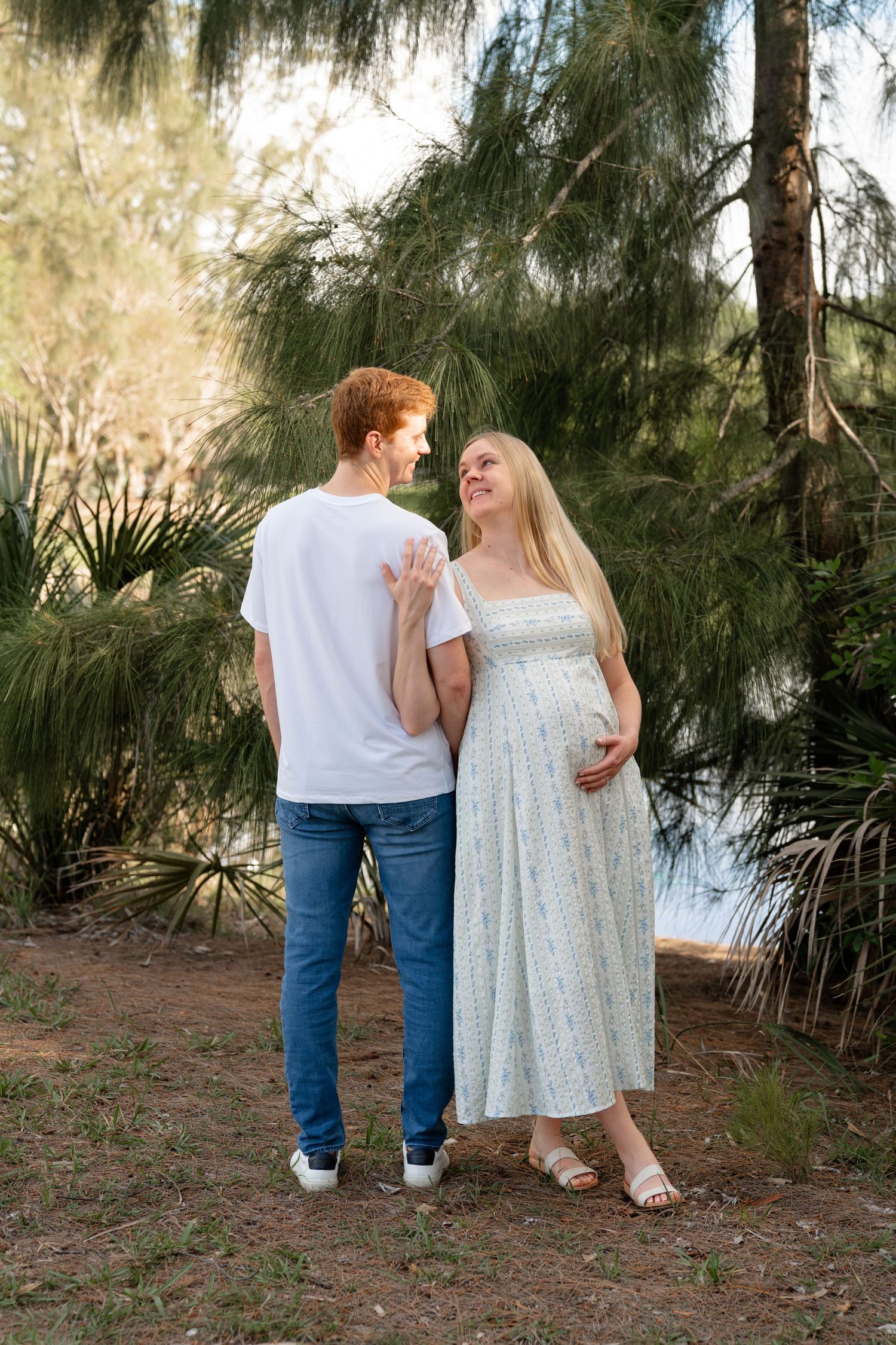 A pregnant woman in a floral dress stands with a redheaded man in a park, both smiling at each other near pine trees.