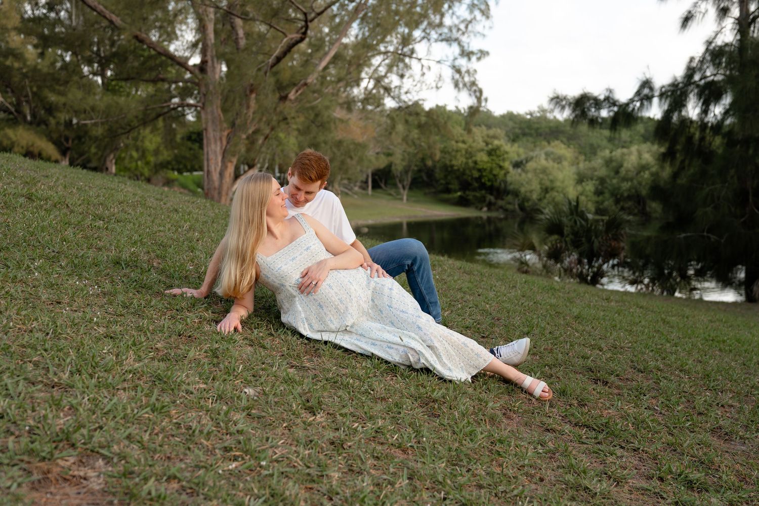 Couple sitting on grass by a pond, woman in white dress leaning on man in jeans, surrounded by trees.