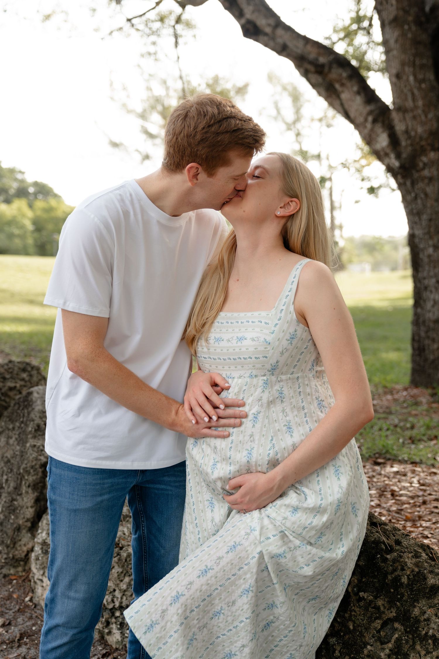 Pregnant couple sharing a tender kiss outdoors, hands on baby bump, wearing casual white top and floral dress.