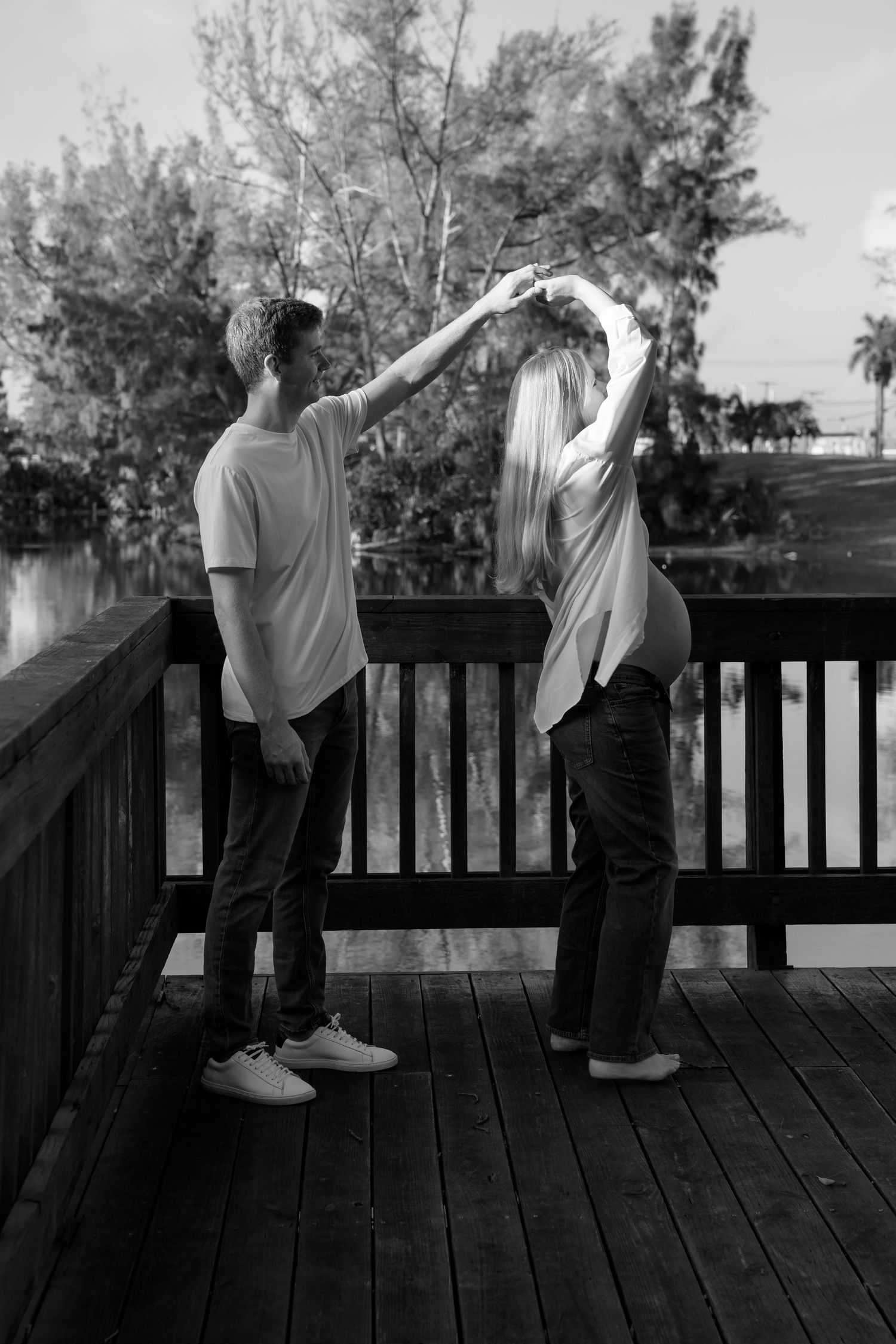 Black and white photo of a couple dancing on a wooden deck near a lake, holding hands raised above their heads.