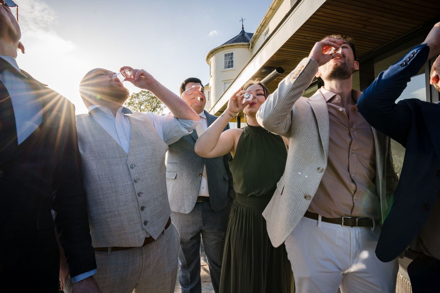 Elegantly dressed wedding guests laughing and toasting drinks outdoors in golden sunlight near a historic building.