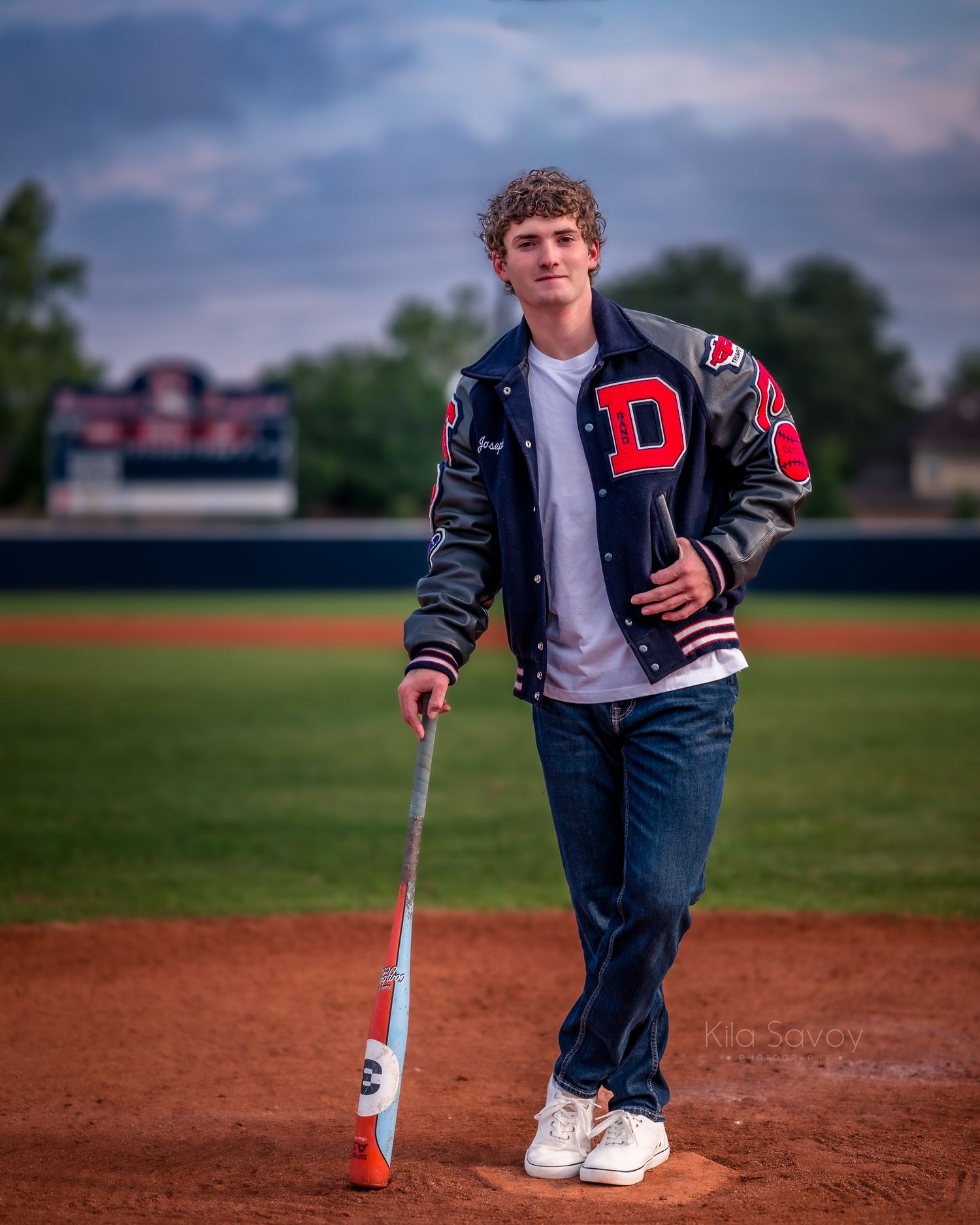 Teen baseball player in varsity jacket tossing a ball while leaning on a bat near a brick wall.