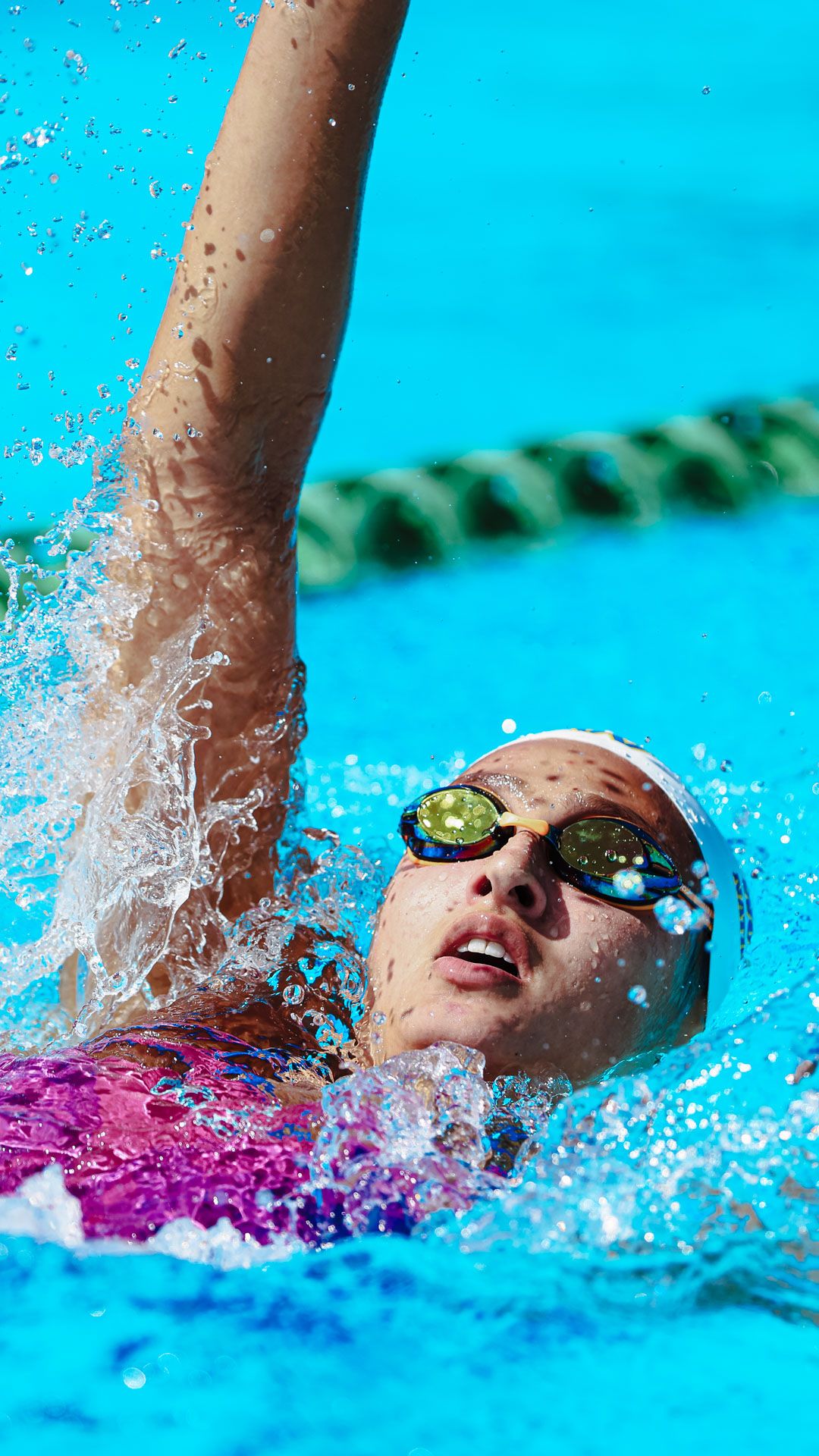 Female swimmer doing backstroke in pool, wearing goggles and swim cap, water splashing around her.