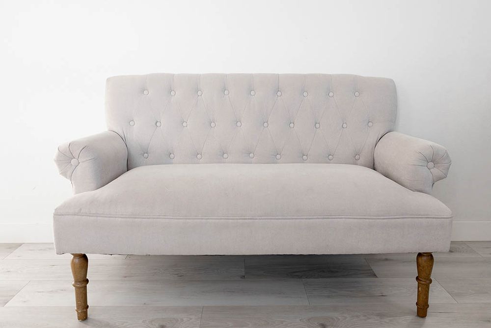 Beige tufted loveseat sofa with rolled arms and wooden legs on light floor.