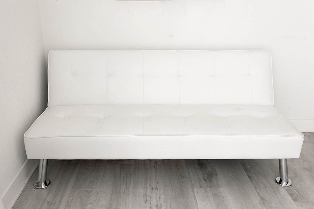 White faux leather futon sofa bed with chrome legs on light hardwood floor.