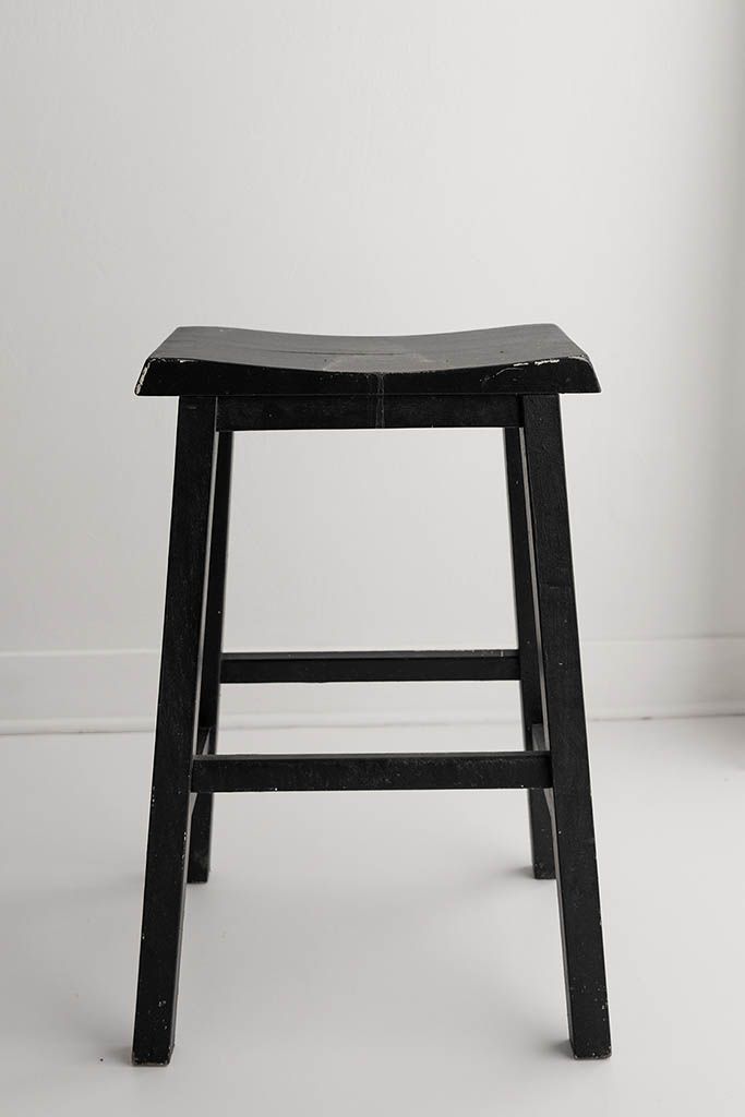 Black wooden saddle seat bar stool with four legs on white floor.