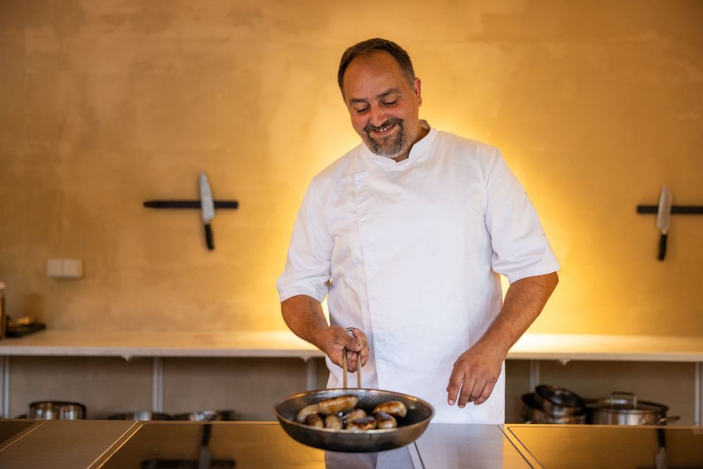Chef in white uniform smiling while cooking mushrooms in a pan in a modern kitchen.