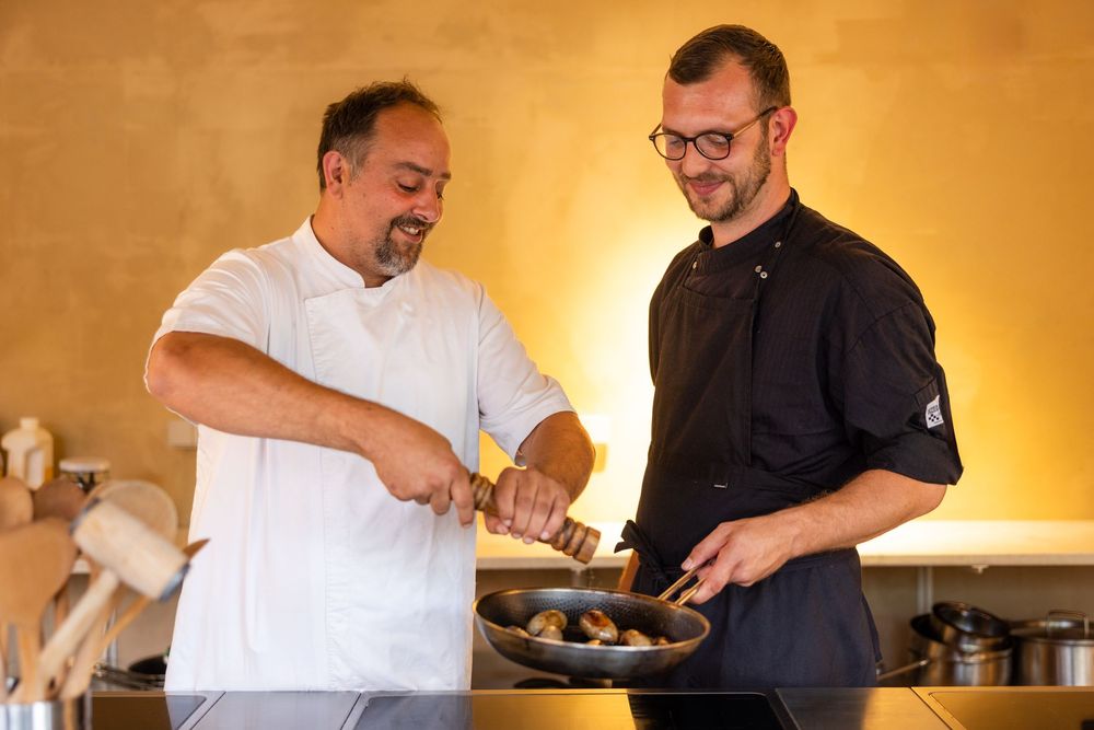 Two chefs in a professional kitchen smiling while sautéing food in a pan together.