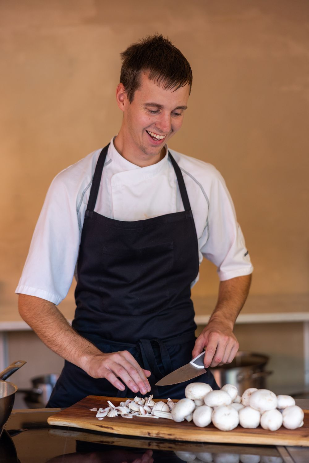 Smiling chef in black apron chopping white mushrooms on wooden cutting board in professional kitchen.