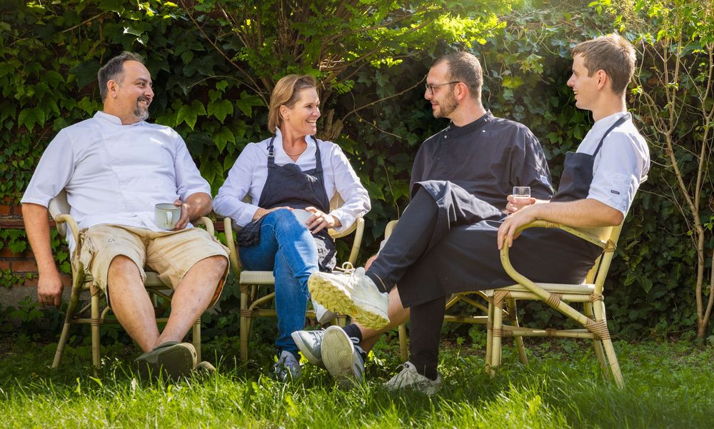Four restaurant staff in aprons relaxing on wicker chairs in a sunny green garden, enjoying a casual conversation outdoors.