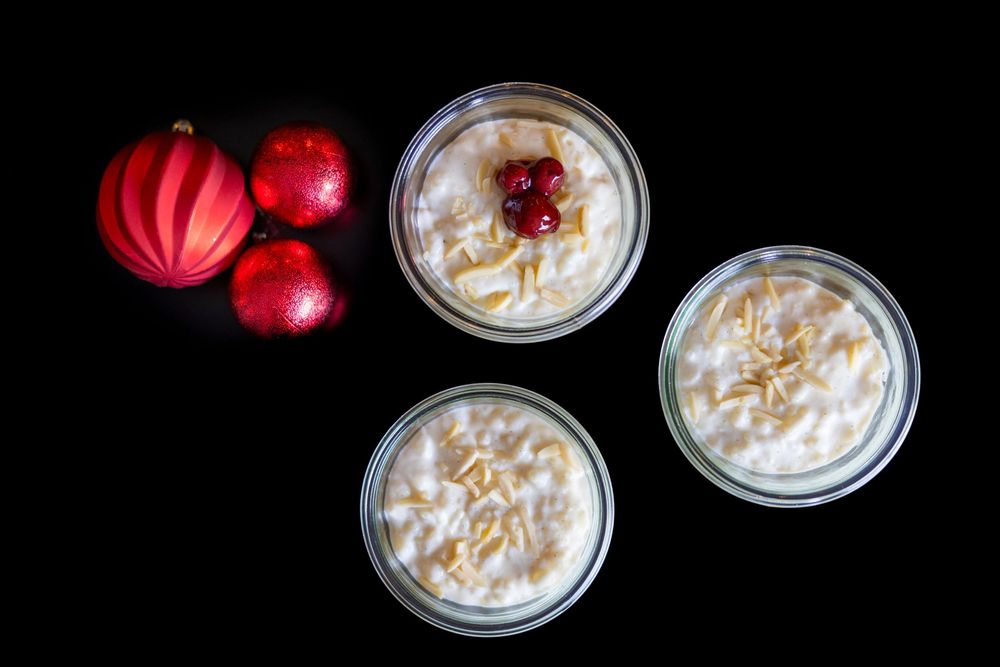 Three glass jars of creamy rice pudding topped with almonds and cherries, displayed with red Christmas ornaments on black background.