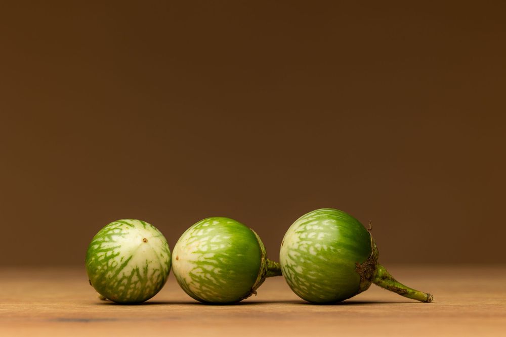 Three small green striped eggplants arranged in a row on a wooden surface against a brown background.