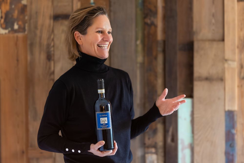 Woman in black turtleneck holding a wine bottle, presenting in a rustic wooden interior setting.