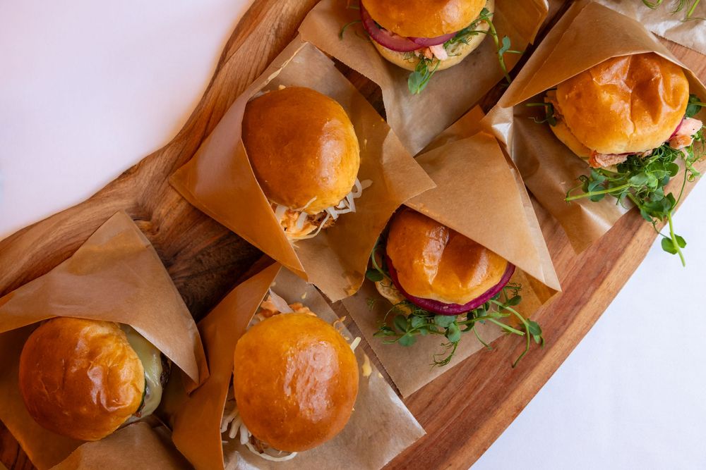 Six gourmet sliders wrapped in parchment paper on a wooden board, topped with fresh microgreens and colorful toppings.