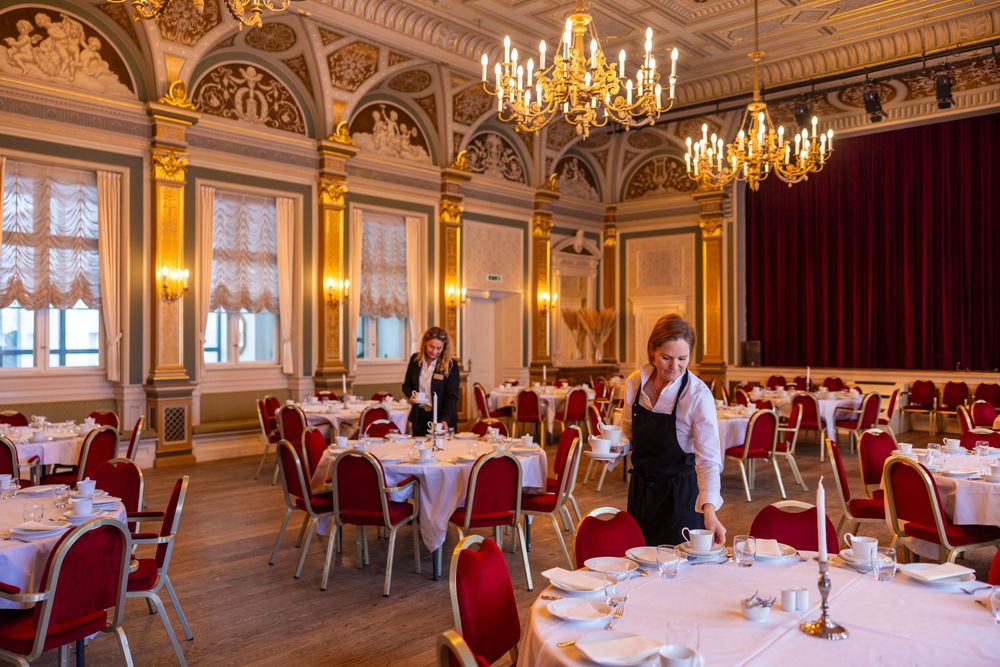 Elegant banquet hall with white-clothed tables, red chairs, gold chandeliers, and staff preparing for a formal dining event.