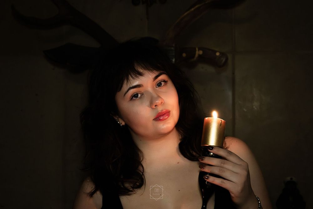 Young woman with dark curly hair holds a lit candle in dim atmospheric lighting with antlers visible in background.