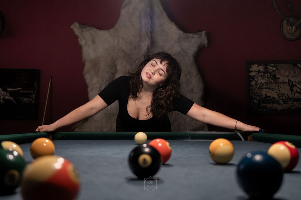 Woman leaning over a pool table with billiard balls in foreground, in a dark red room with animal hide on wall.