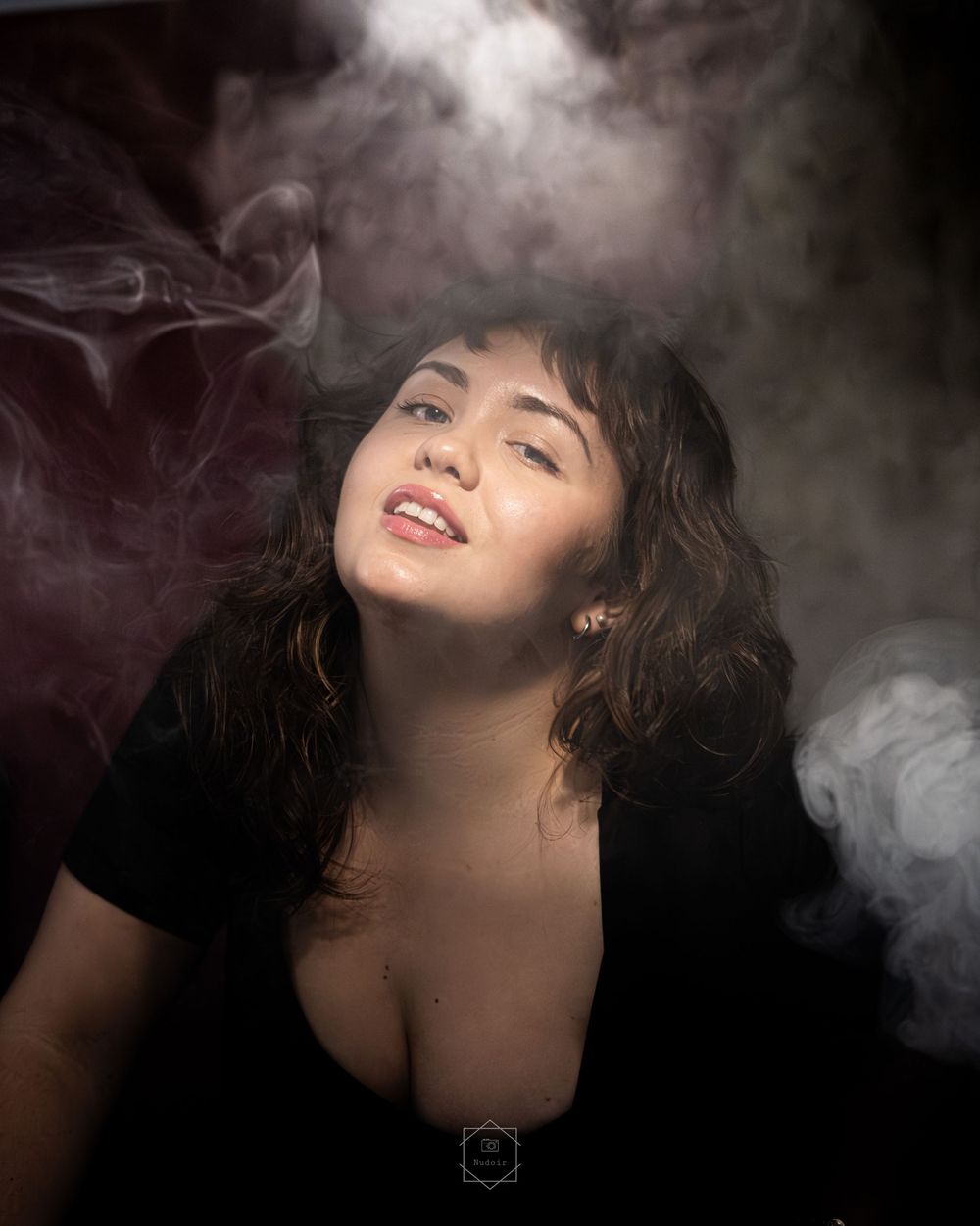 A young woman with curly dark hair poses dramatically surrounded by swirling smoke clouds against a dark studio background.
