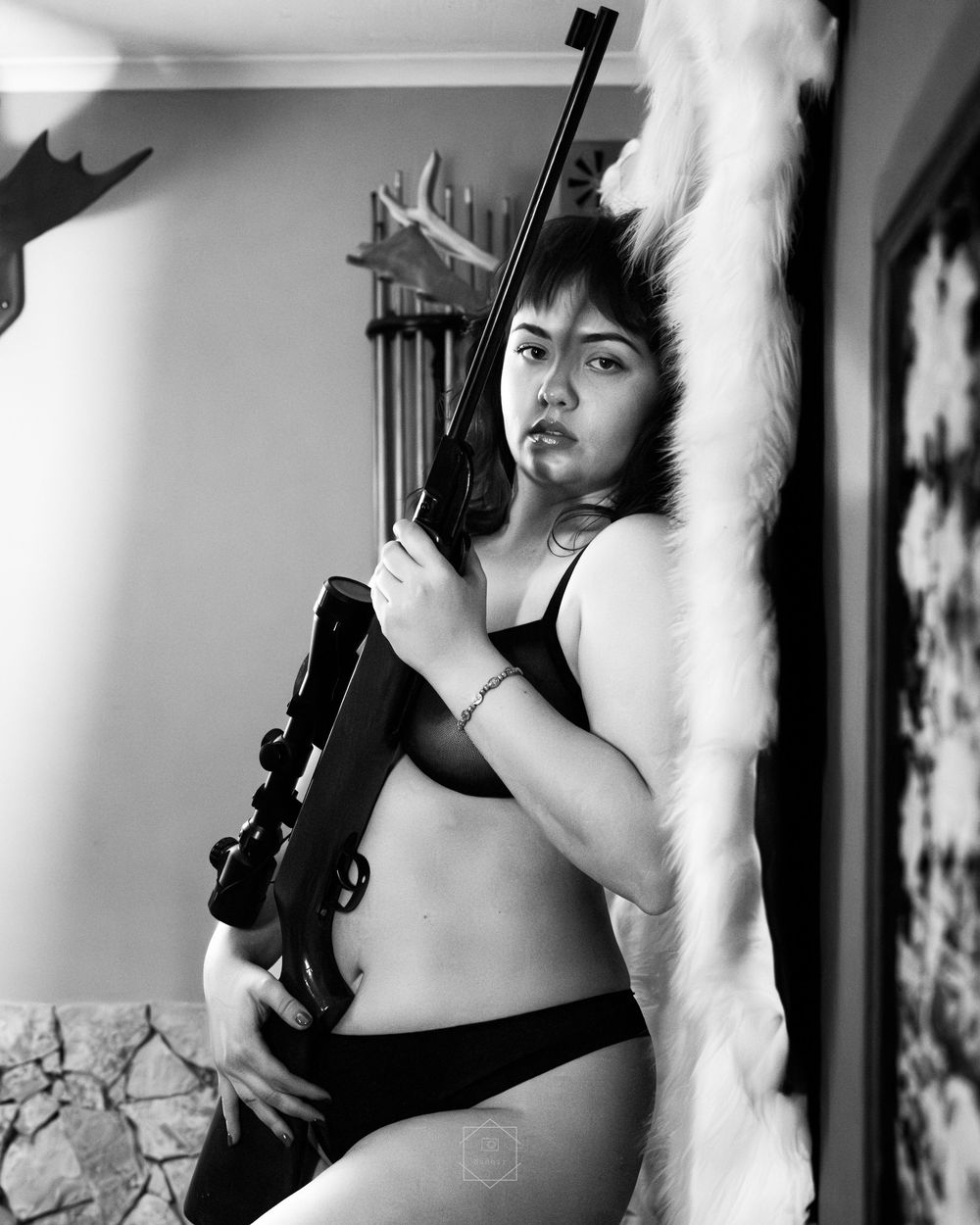 Black and white photo of a woman in lingerie holding a rifle while leaning against a fur-draped wall indoors.