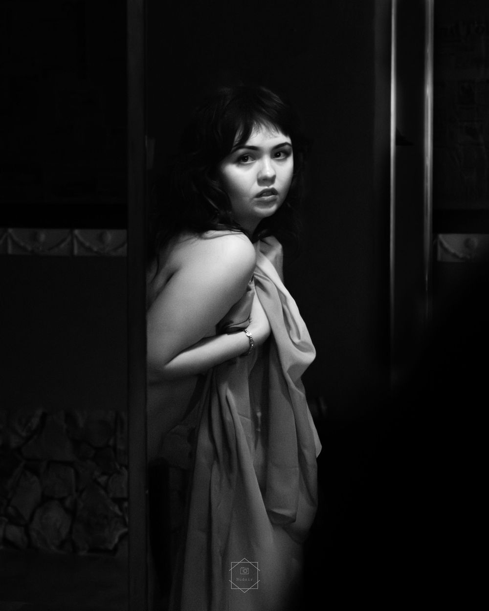 Black and white portrait of a young woman wrapped in sheer fabric, posing dramatically in a dark doorway.