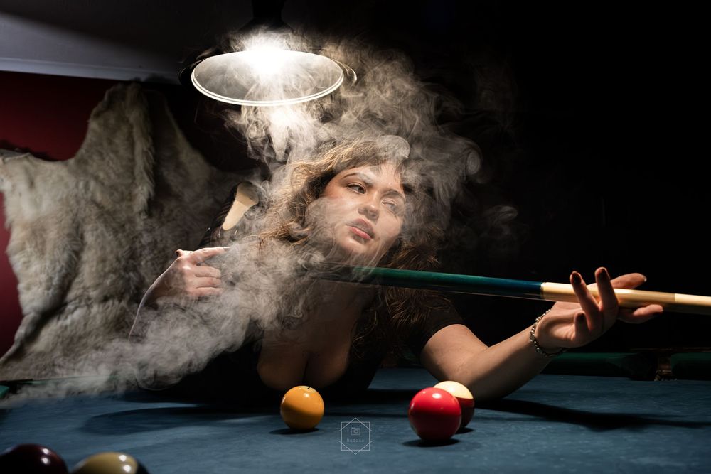 Woman playing pool surrounded by dramatic smoke under spotlight in dark billiards room with colorful balls.