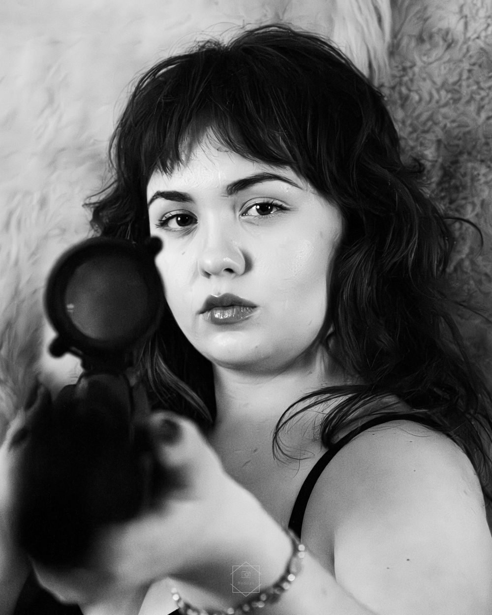 Black and white portrait of a young woman with dark wavy hair holding a camera lens toward the viewer.