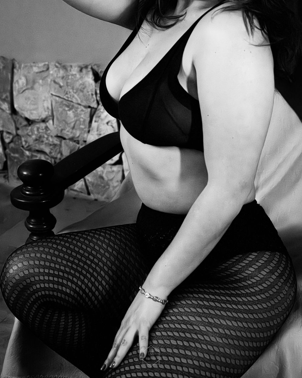Black and white photo of a woman in a black bra and fishnet tights posing elegantly on a bed.