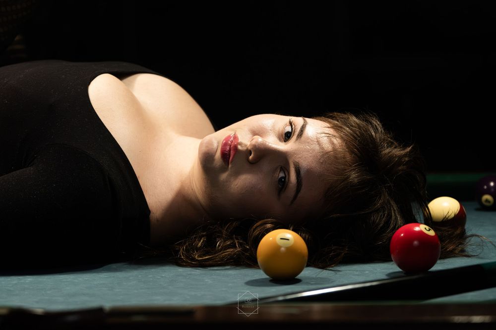 Woman lying on a pool table surrounded by billiard balls, wearing black top, dramatic lighting.