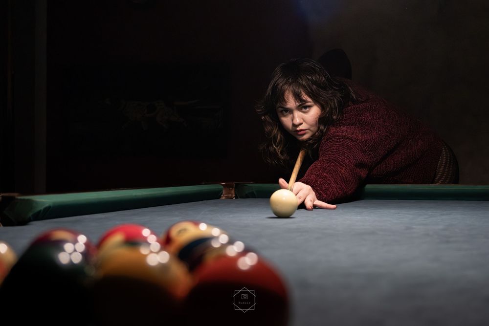 Woman in red sweater leaning over a pool table, aiming cue stick at billiard balls in a dimly lit room.