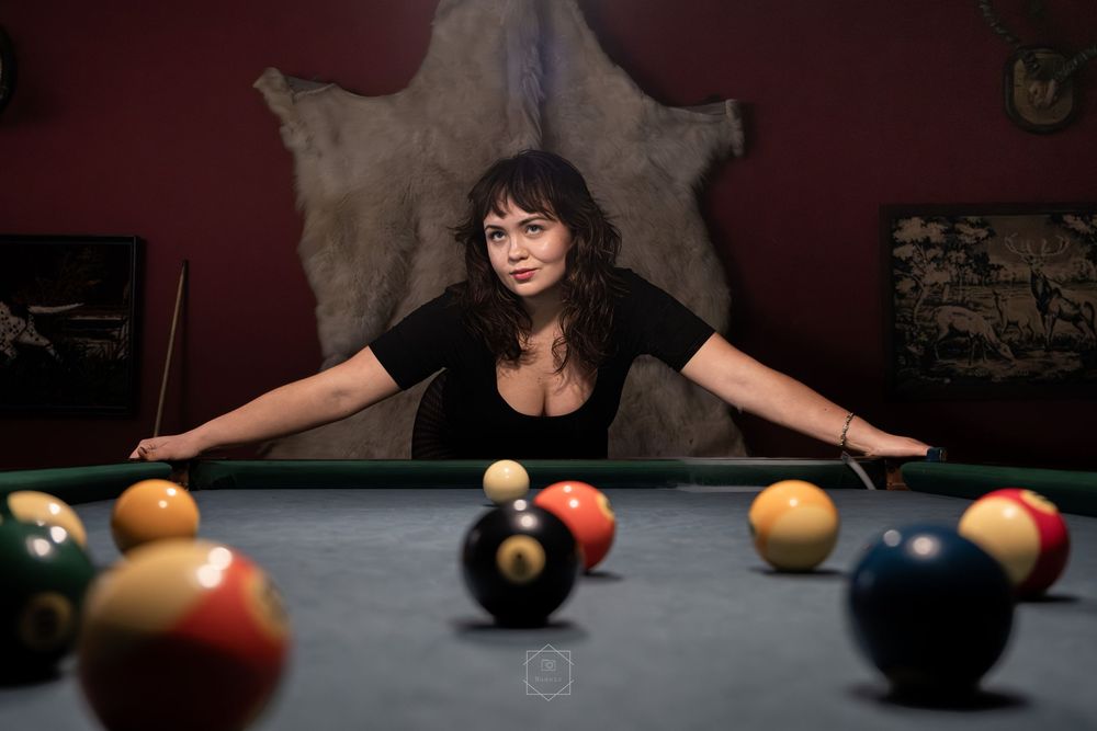 Woman leaning over a pool table surrounded by billiard balls in a dark, moody game room.