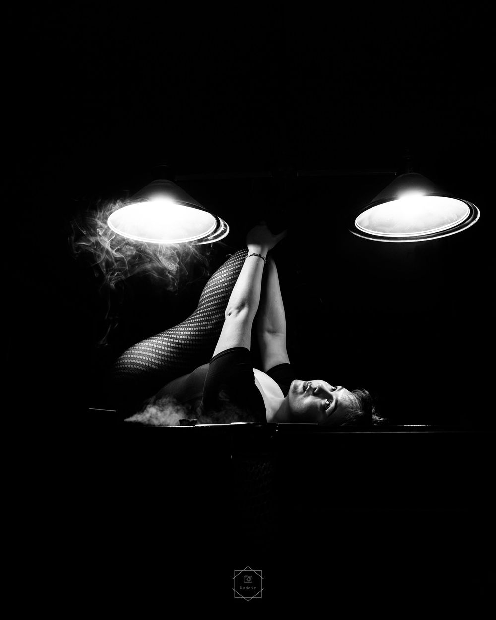 Black and white photo of a woman lying down with legs raised between two glowing lights in a dark room.